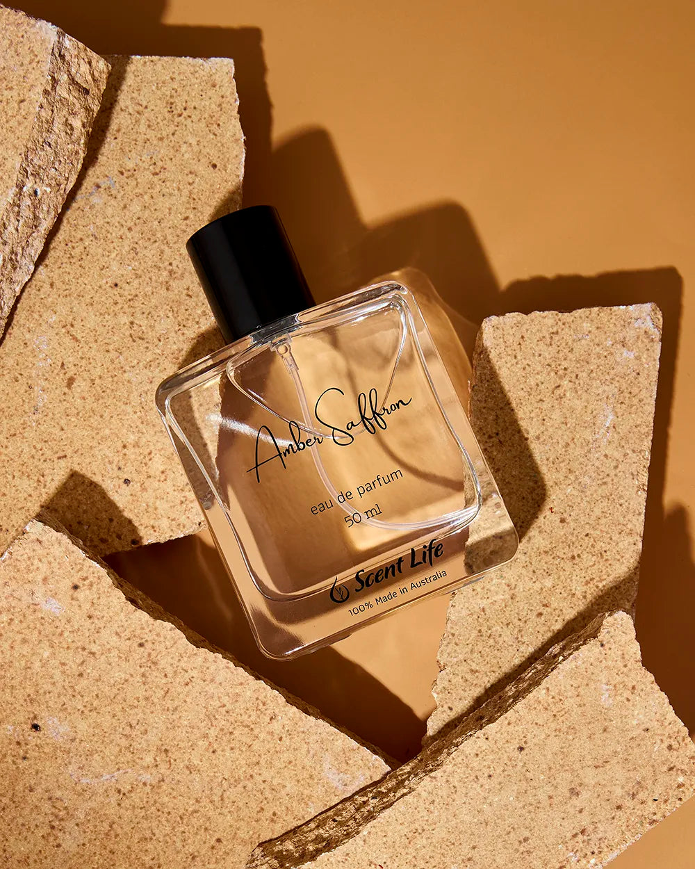 Bottle of Amber Saffron perfume lying diagonally on brown coloured stones with an amber background. The bottle is square, transparent, with a black lid and cursive font.