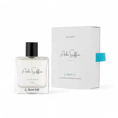 An Amber Saffron Eau De Parfum glass bottle from Scent Life, featuring a minimalist design and a black cap, is showcased next to its white box packaging. The elegant typography and blue ribbon accentuate the allure of this captivating fragrance.