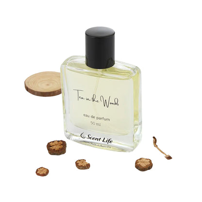 A 50 ml Eau de Parfum bottle of Scent Life's Tea in the Woods, renowned for its long-lasting fragrance, is displayed in a clear glass bottle with a sleek black cap. It elegantly sits next to small wooden slices against a pristine white background.